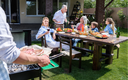 Abgeschnittene Aufnahme eines Mannes, der ein Tablett mit Fleisch hält, während die Familie im Freien am Tisch sitzt.png
