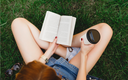 Die Frau trägt blaue Shorts, sitzt mit einer Tasse Kaffee auf einem grünen Gras und liest das Buch aus der Nähe.png