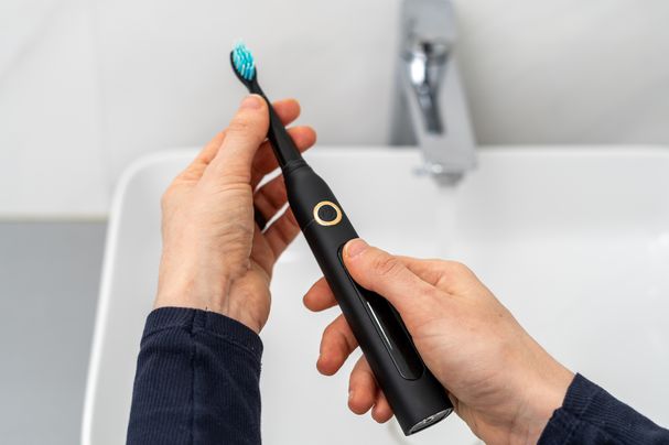Die Pov-Frau hält professionelle akustische elektrische Zahnbürste mit austauschbarer Bürste in den Händen über dem Waschbecken auf unscharfem Hintergrund. Drahtlose Geräte und Mundpflege.jpg