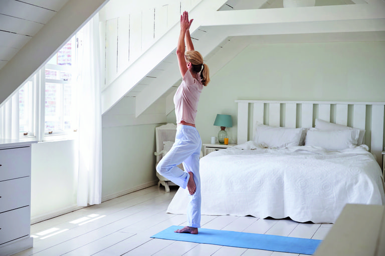 frau in yogapose steht im schlafzimmer und macht yoga mit fensterausblick