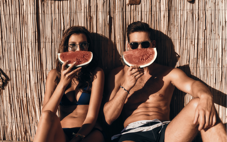 Schönes junges Paar, das eine Hälfte seiner Gesichter mit einer Scheibe Wassermelone bedeckt, während es im Freien sitzt.png