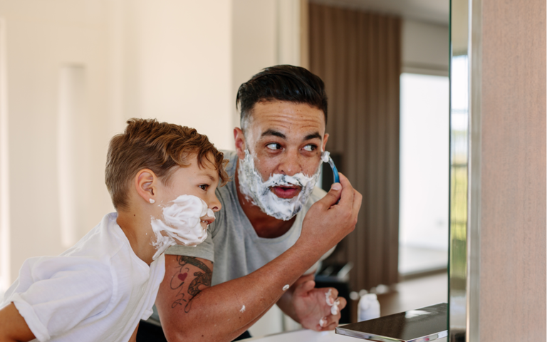 Vater und Sohn rasieren sich im eigenen Badezimmer zusammen. Junge Männer und kleine Jungen mit Rasierschaum auf ihren Gesichtern rasieren und schauen in den Spiegel..png