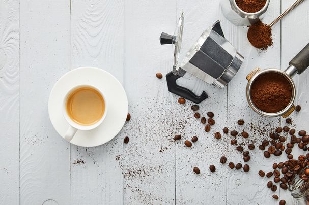 eine tasse kaffee gemahlener kaffee und ein aufgeschraubte espressokocher inlusive kaffeebohnen auf einem holztisch ausgestreut
