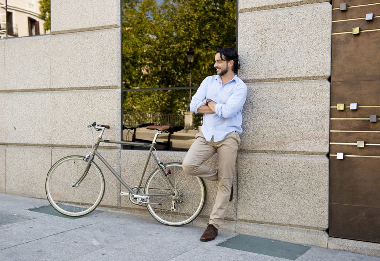 lächelnder Mann in blauem Hemd und beiger Hose lehnt an Hausfassade mit verschränkten Armen und hat ein Fahrrad neben sich stehen.jpg