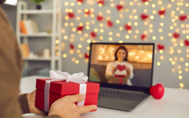 mann-überrascht-freundin-am-laptop-mit-geschenk-mit-valentinstagsdeko