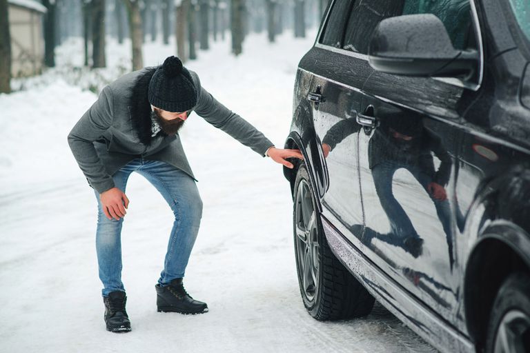 mann in mantel und mit haube kontrolliert den hinterreifen eines autos bei winterlichem wetter